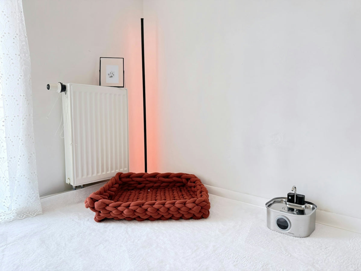 A chunky knit rust-colored pet bed on a white floor near a radiator
